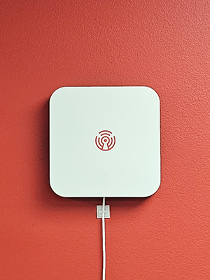 The Room Alert MAX Base Station, a white, square-shaped device with rounded corners and the Room Alert logo, mounted on a red wall with a visible white power cord.
